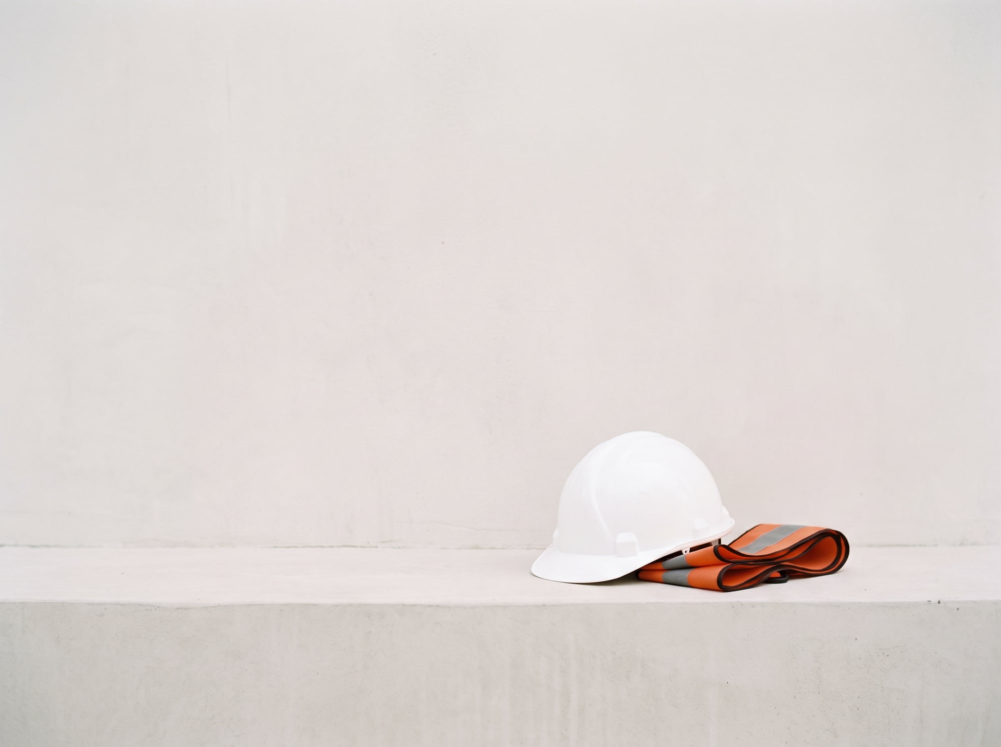 A white hard hat and folded orange high-vis strap on a concrete ledge