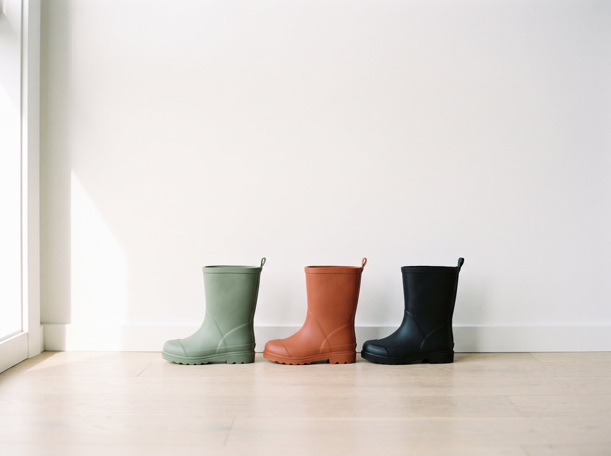Three small gumboots in sage, terracotta and black on a pale timber floor
