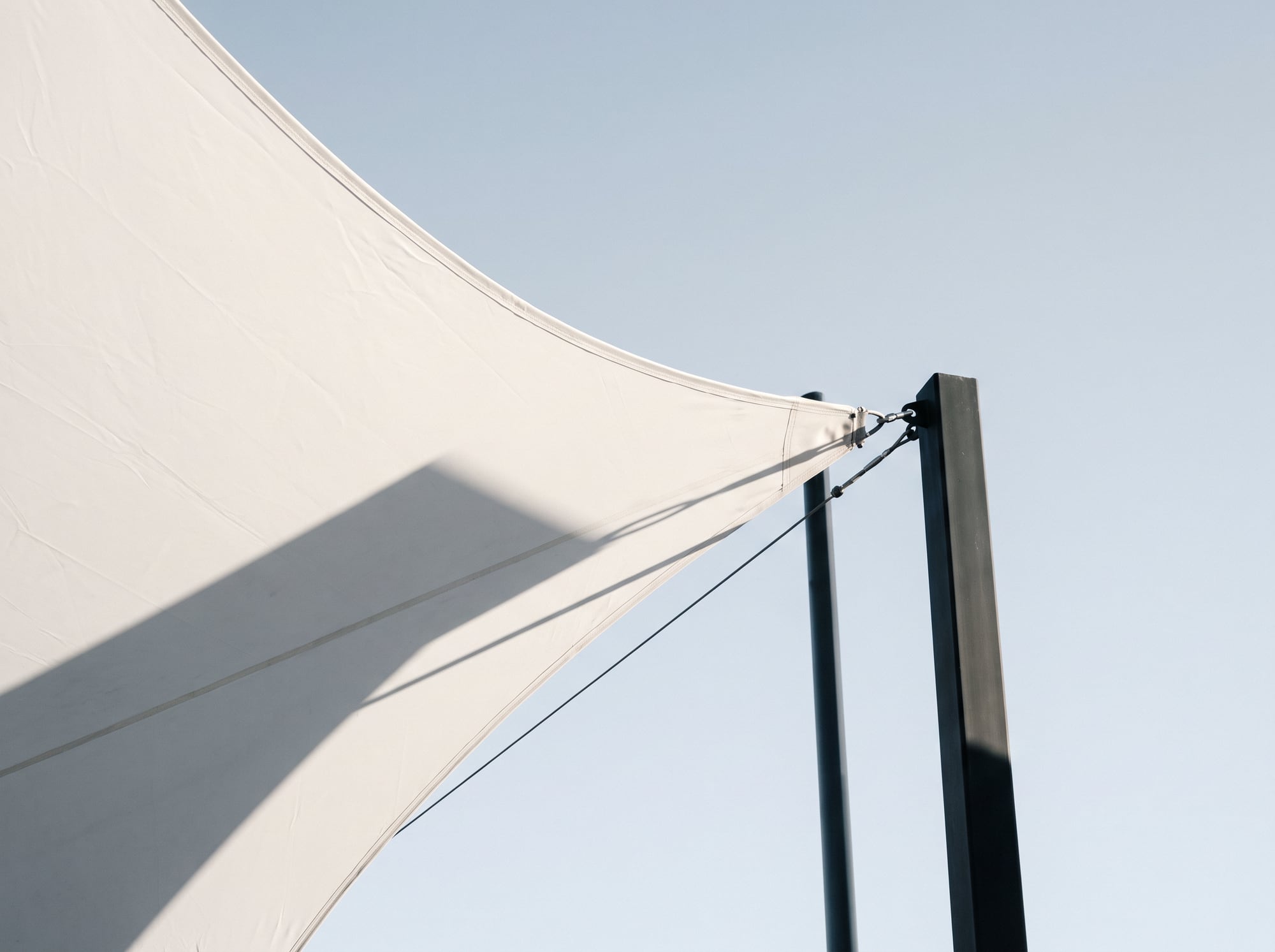 A white shade sail and black steel post against a pale sky